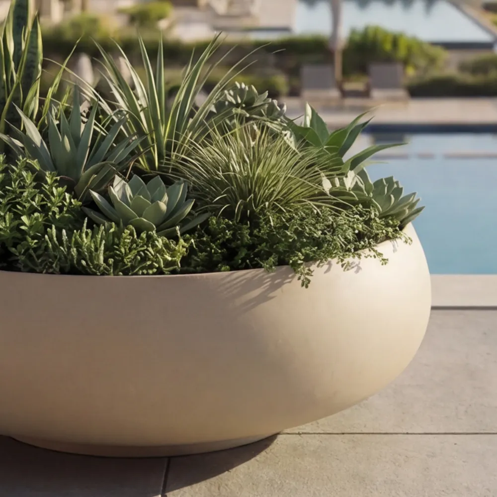 Detalhe da floreira Ariel com corpo arredondado bege e composição densa de folhagens e suculentas junto à piscina.