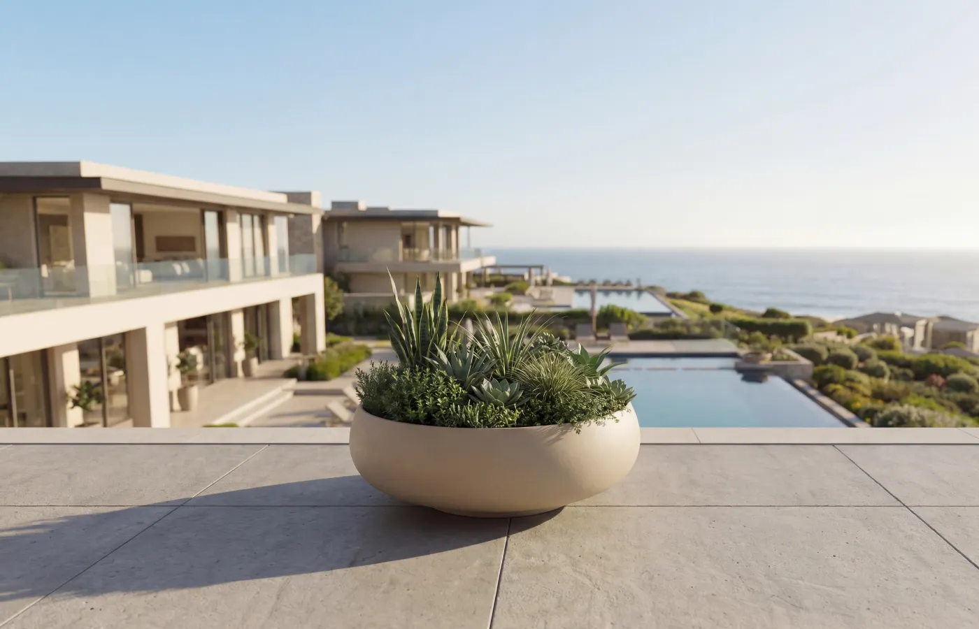 Floreira Ariel em terraço com vista mar, com corpo bege arredondado baixo e composição densa de folhagens.
