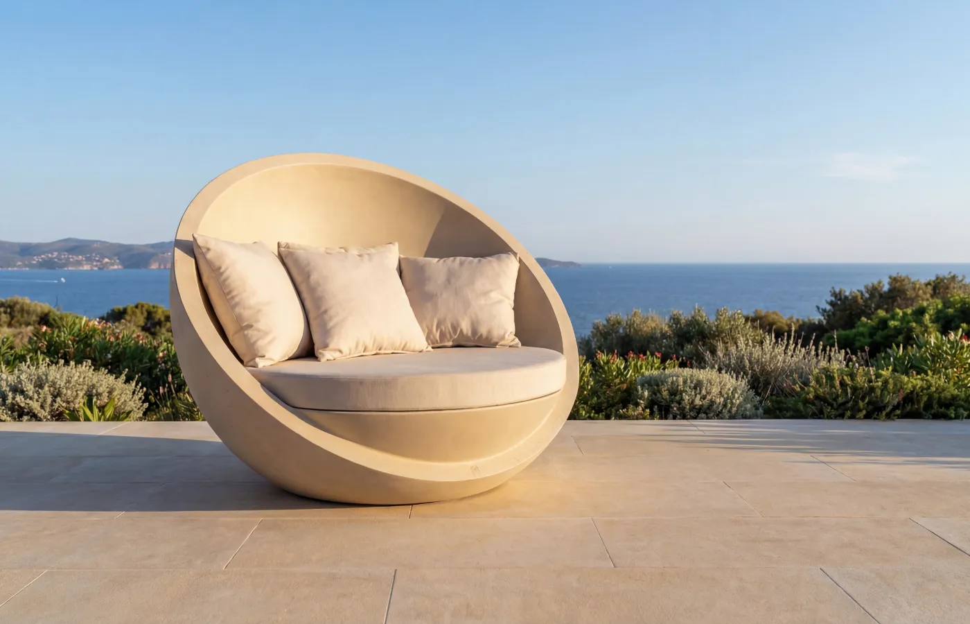 Bola outdoor sofa in beige on a terrace with sea view, showing its enveloping circular shape and cushioned seat arrangement.