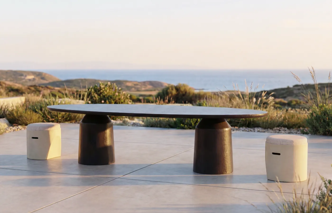 Siana oval outdoor dining table in brown, with two cylindrical bases, set between two Cloe stool with a sea view.
